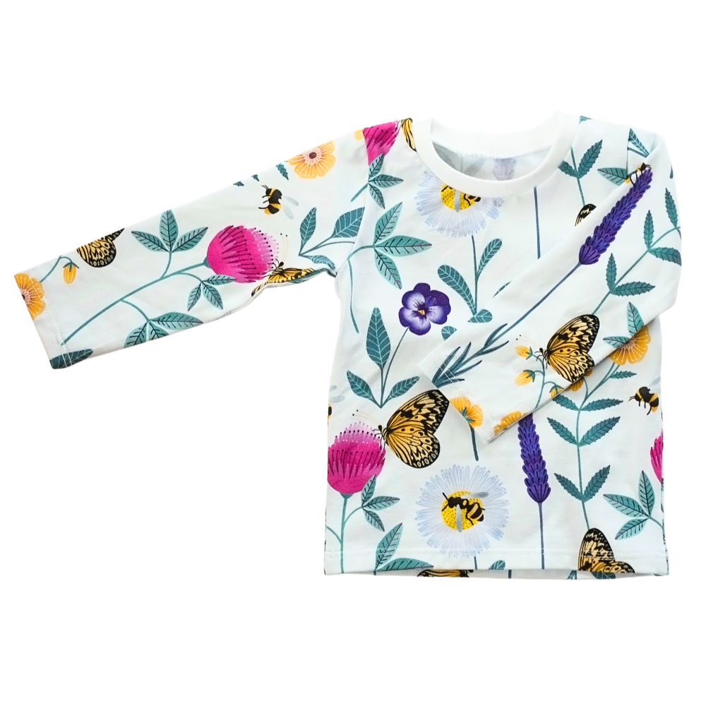 Platt liggande långärmad trikåtröja i vit ekologisk bomullsjersey med färgglatt mönster av blommor och insekter i lila, rosa, gult och turkos.